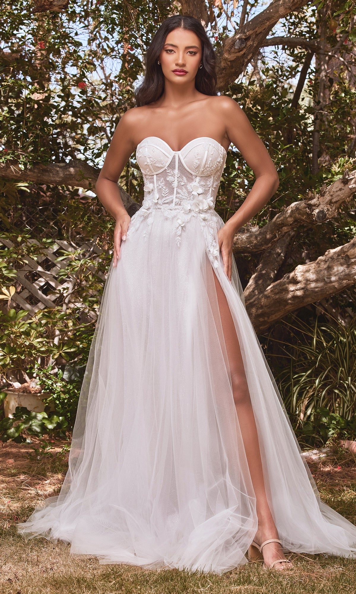 A woman stands outdoors among trees in the Puff-Sleeve Off-White Long Prom Dress CB080W, featuring a sheer bodice, floral appliqué, high slit, and sheer puff sleeves.